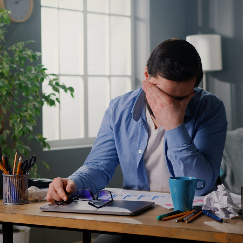 Close up of young man sitting at workplace at home office. Tired businessman rubbing eyes, feeling exhausted. Daily routine.